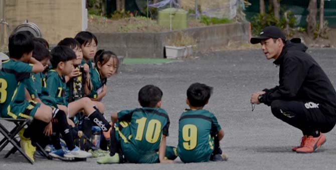 コーチが座りながら子供たちに指導している写真